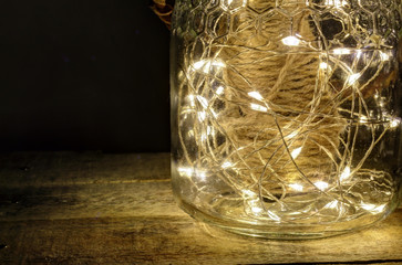 A still life composition with a glass jar with a leather cover filled with a hemp rope and some bright shining leds on a rought wood