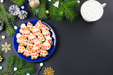 Tasty Christmas cookies, hot drink with marshmallows and decorations with Christmas tree and lights on black background.