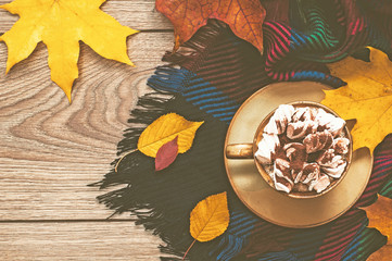Fototapeta na wymiar A cup of coffee with marshmallows and chocolate and a warm scarf on a wooden surface. Yellow leaves, autumn background. Top view