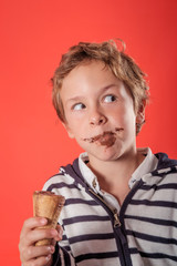 close up of one young blond and cute boy eating and holding with his hand one chocolate ice-cream over a red background in studio. He has a chocolate stains on his face and a mischievous look.