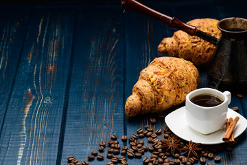 coffee pours into a Cup on a blue wooden table with coffee beans and a croissant