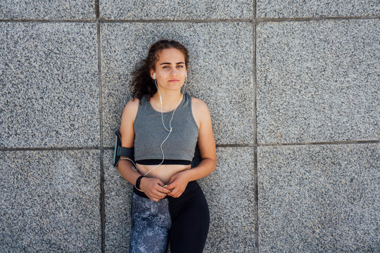 Portrait of young athletic woman wearing earbuds leaning against a wall