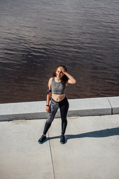 Exhausted young athletic woman wearing earbuds standing at the riverside