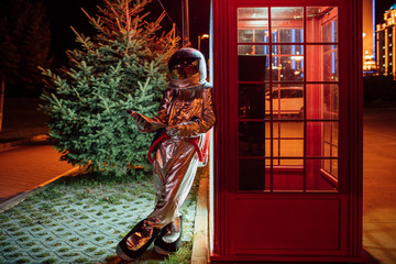 Spaceman leaning against a telephone box at night reading phone book