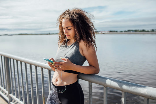 Young athletic woman wearing earbuds and using smartphone at the riverside