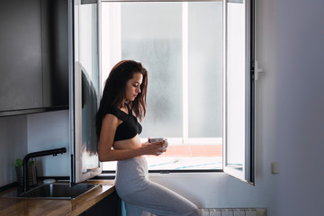 Fototapeta na wymiar Serious beautiful young woman with cup of coffee at the window at home