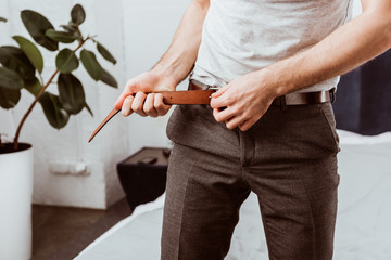 partial view of businessman putting on belt in bedroom at home