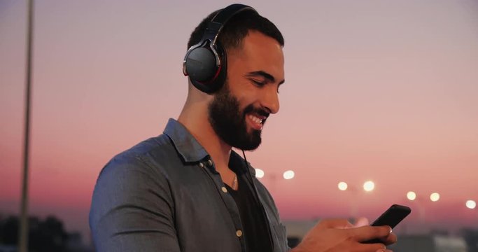 Handsome Bearded Man Walking in the Night City. Parking full of Cars in the Background. Man Holding Mobile Phone. Listening to Music. Having Fun. Big Headphones on his Head.