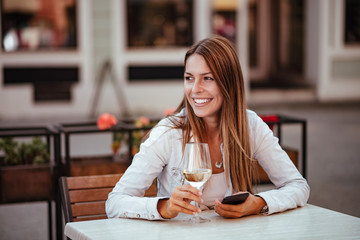 Portrait of a smiling brunette woman having a glass of wine at restaurant outdoors. Holding smartphone and looking away.