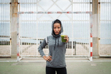 Confident urban sporty woman offering green detox smoothie. Successful fitness nutrition and outdoor workout concept.