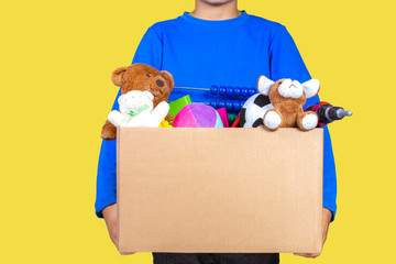 Donation concept. Kid holding donate box with clothes, books, school supplies and toys, yellow color background