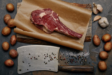 flat lay with raw rib eye steak on baking paper with butcher knife, spices, potatoes and garlic