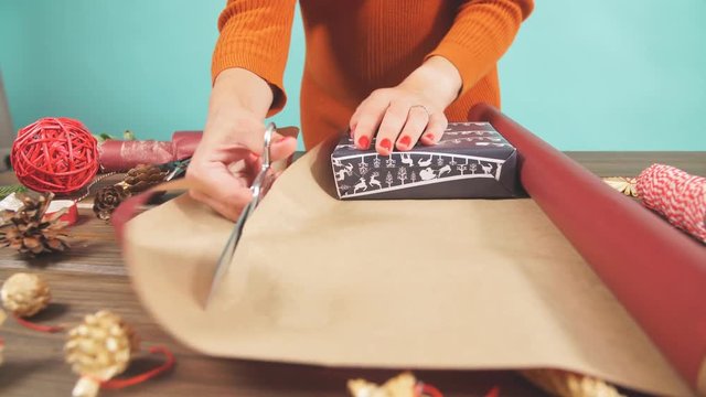 Woman in a red sweater wrapping gift box with equipment and decorating items on wood table, preparing for celebrating Christmas holidays - top view.