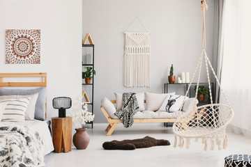 Bright living room interior with macrame on the wall, beige couch with pillow and blanket, hammock chair, fluffy rug and bedside table with lamp standing by the bed in the real photo