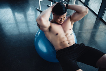 focused young asian sportsman with bare chest doing abs exercise on fitness ball at gym