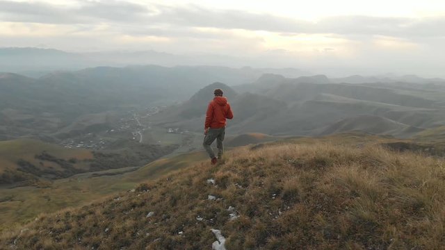 An aerial view of a bearded hipster male with a remote from a drone walks along the edge of a high plateau near a cliff at sunset. The guy is on the cliff
