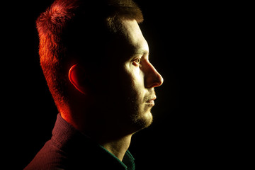 Close-up of the profile of an unshaven face of a man with stubble dressed in a shirt in the dark, illuminated from the front by a yellow back red on a black isolated background