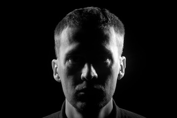 Monochrome close-up face of the unshaven man with bristles dressed in a shirt in the dark, highlighted on the left and right side and in the middle of the shadow on a black isolated background