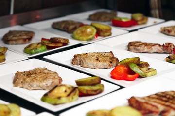 Many white ceramic square plates with grilled meat steaks and colored grilled vegetables close-up. Banquet concept in restaurant, birthday