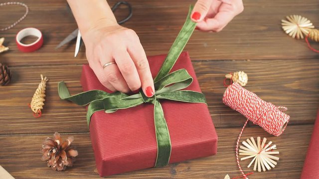 Close-up top view of female hands decorating red gift boxes with backer s twine and fir brunches on wooden table. Pine cones and christmas decoration on dark desk.