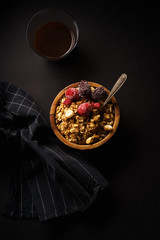 Granola with raspberries and blackberries in wooden bowl with a cup of coffee on black table.