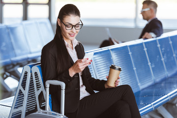 young smiling businesswoman in glasses with coffee to go using smartphone in departure lounge at...