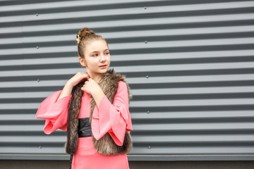 closeup portrait of cute smiling little girl in pink dress and fur sleeveless jacket posing near gray corrugated striped wall