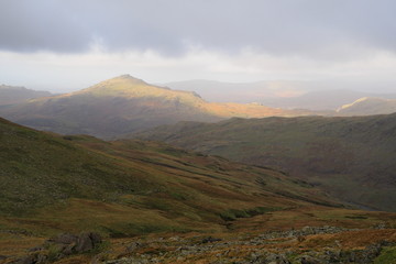Obraz premium Sunlight falls on a Lake District mountain top out of an overcast and cloudy sky lighting it up with warm Autumn colours, Cumbria, UK