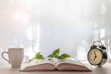 An open book, a gray cup with coffee, a black clock and a twig with green leaves on a wooden table lit by the sun. Copy space