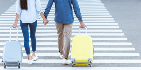 cropped view of couple of tourists crossing pedestrian, holding hands and pulling their luggage