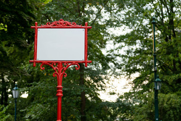 Red decorative metal frame with white banner for text. In the background trees with green leaves.