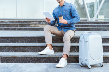 Man sitting on the staircase with luggage, holding digital tablet and coffee to go