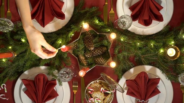 Female hand lights christmas candles. Festive red table setting with garland. Top view