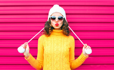 Funny girl blowing red lips makes air kiss wearing colorful knitted yellow sweater hat over pink...