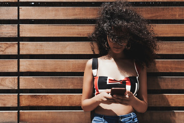 A young caucasian female in the glasses is leaning against a wooden striped wall and typing a...