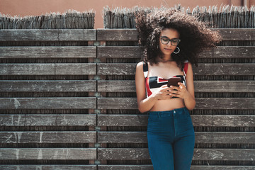 A cheerful young biracial female is leaning against a wooden fence with wattle inside, laughing, and holding the cell phone, with a copy space area on the right for text, your logo or advertising