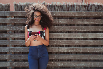 A cheerful young biracial female is leaning against a wooden fence with wattle inside, laughing, and holding the cell phone, with a copy space area on the right for text, your logo or advertising