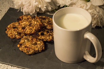 Homemade cookies with a banana lie on a stone board, there is a white cup with milk, in the background white peonies