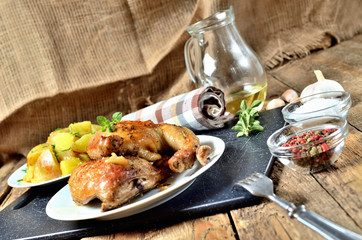 Golden grilled chicken legs on a plate and roasted potatoes, rosemary, jug with oil, oregano, salt, pepper and garlic on a wooden table
