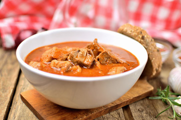 Close-up of pork goulash with pieces of meat in a bowl, garlic, pepper, onion, jug with oil and red checkered tablecloth in the background