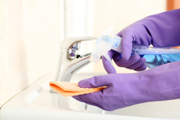 Hands in gloves with sponge and bottle of detergent cleaning water tap in bathroom