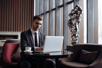 Young handsome man sitting in office with cup of coffee and working on project connected with modern cyber technologies. Businessman with notebook trying to keep deadline in digital marketing sphere.