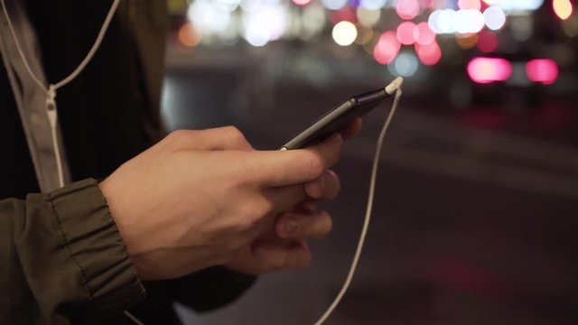 Close-up smartphone hands picking music on mobile phone at night in city. Caucasian male smart phone scrolling on app Stockholm Sweden. Blurry city night background.