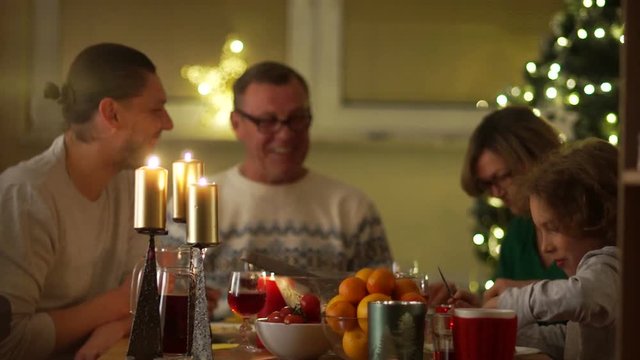 Family Christmas dinner by candlelight on the background of a decorated Christmas tree. A young woman brought roast chicken. Grandfather, grandmother, son and grandson are very happy