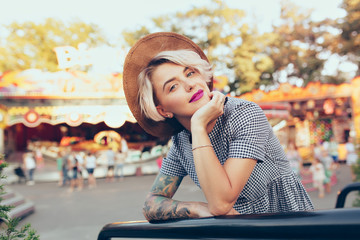 Fototapeta premium Portrait of pretty blonde girl with short haircut posing in amusement park. She wears checkered dress, hat, purple lips and smiles to the camera.