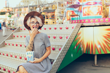 Cute blonde girl with short haircut is sitting on stairs of carousel. She wears checkered dress,  glasses, hat and has purple lips. She holds popcorn and looks funny surprised.