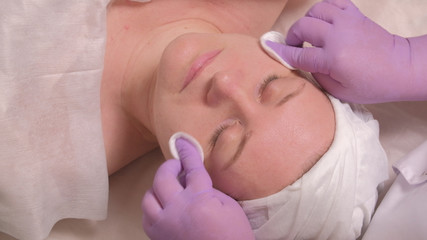A professional cosmetologist in lilac gloves wipes the skin of a female face around the eyes, neck and cheeks using cotton pads. European middle-aged woman on a cosmetic procedure.
