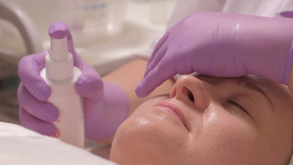 Naklejka premium A professional cosmetologist in lilac gloves with one hand covers his eyes, while the other sprays a moisturizing tonic on the face of a middle-aged woman.