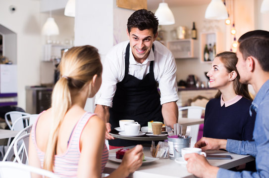 Polite happy waiter bringing ordered dishes to friends in tearoom