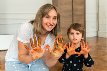 Happy smiling mother and kid playing together, sitting on the floor and painting with tempera...